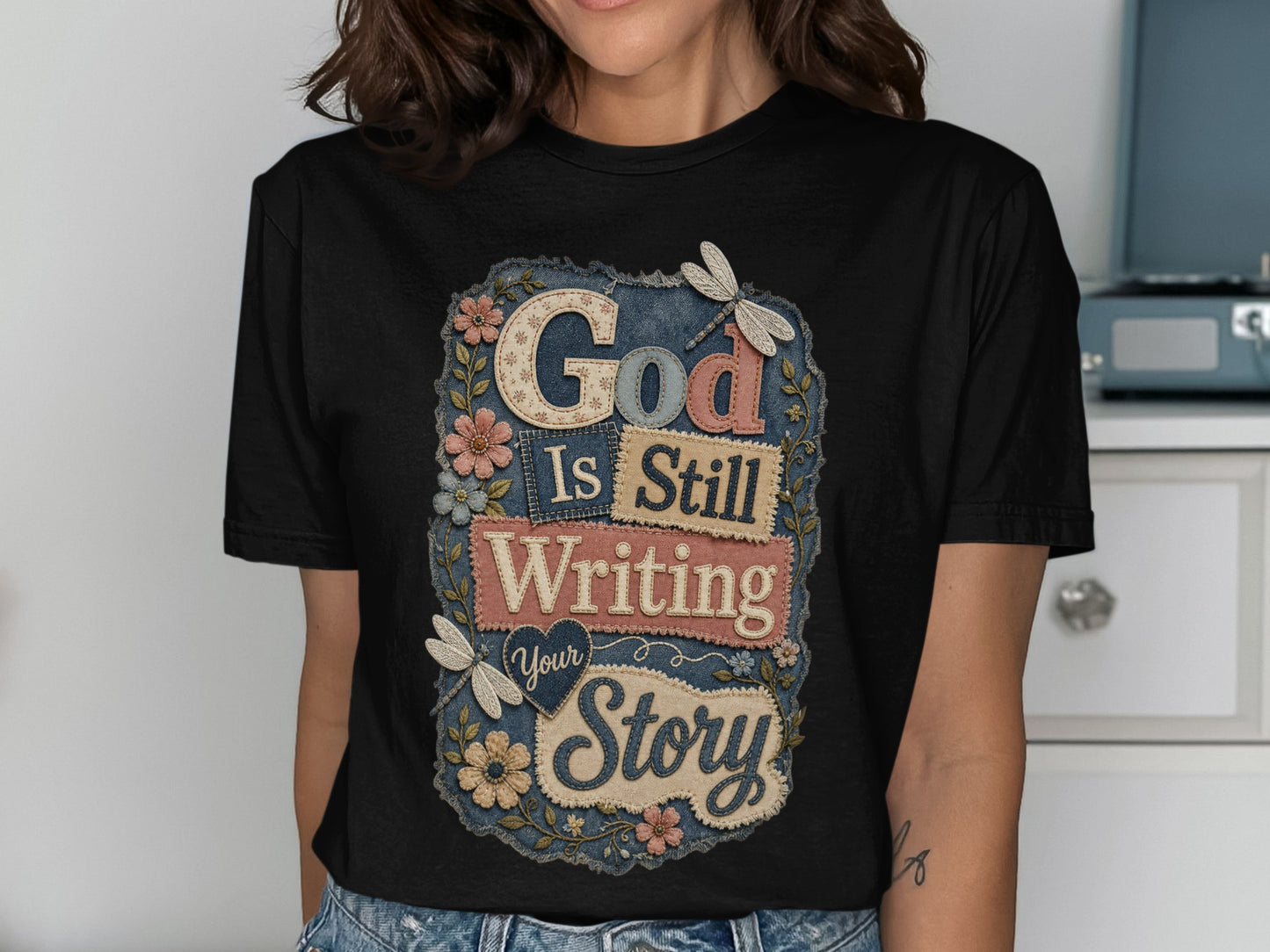 Black t-shirt with "God Is Still Writing Your Story" text and floral accents.