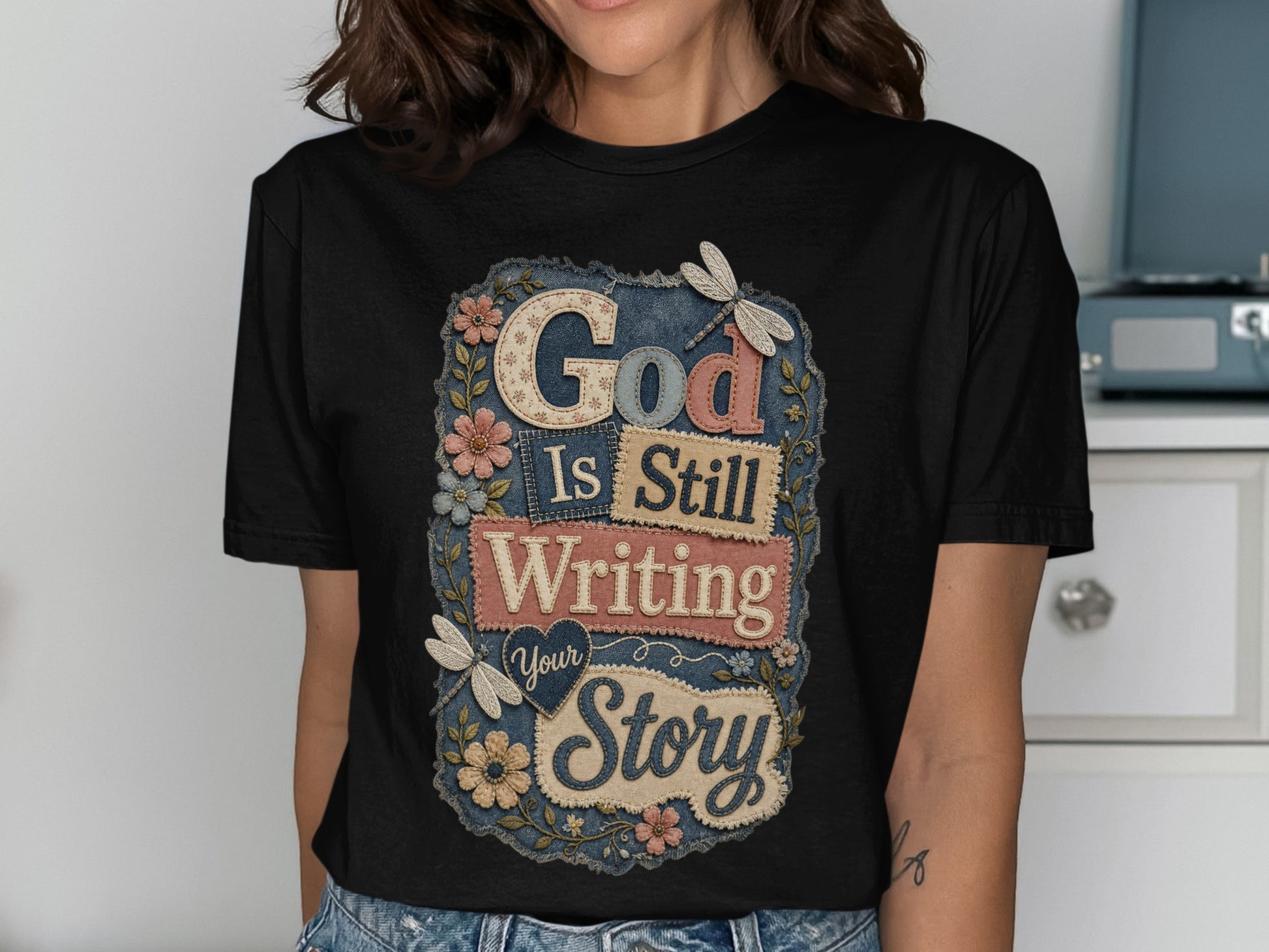 Black t-shirt with "God Is Still Writing Your Story" text and floral accents.