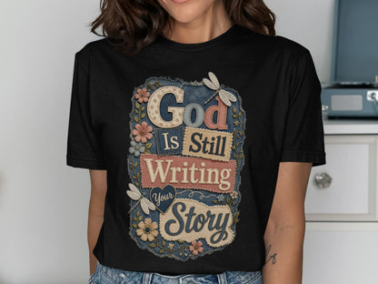 Black t-shirt with "God Is Still Writing Your Story" text and floral accents.