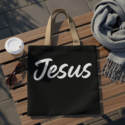 Black tote bag with "Jesus" in white cursive lettering, next to sunglasses and coffee.