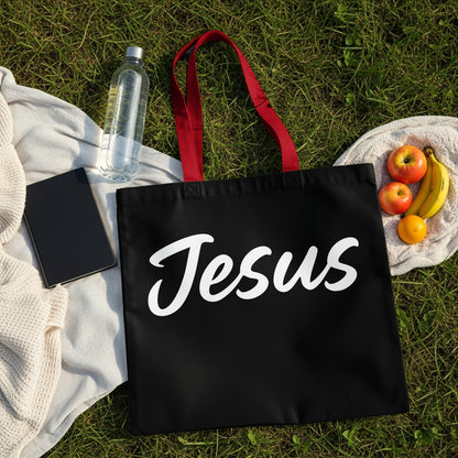 Black tote bag with white cursive "Jesus" text and red straps on grass.