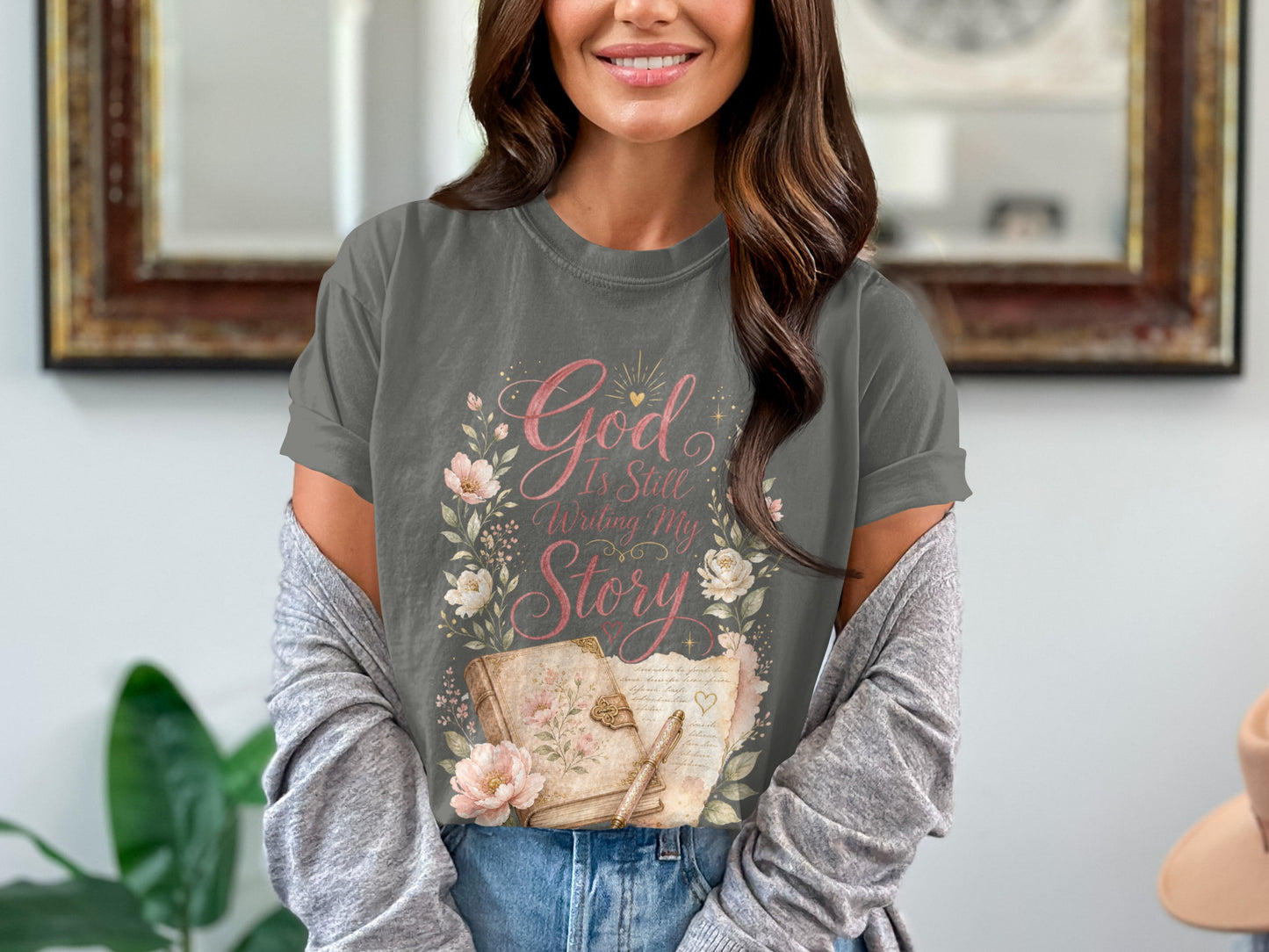 T-shirt graphic: "God Is Still Writing My Story" with flowers, an open book, and a quill pen.