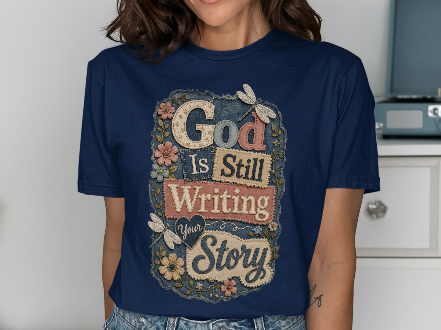 Dark blue t-shirt with "God Is Still Writing Your Story" text and floral accents.
