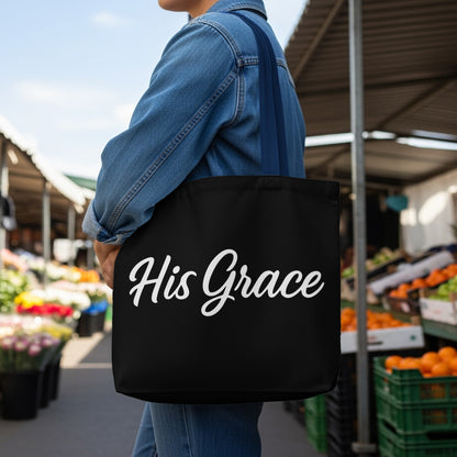 Black tote bag with "His Grace" in white cursive text displayed at an outdoor market.