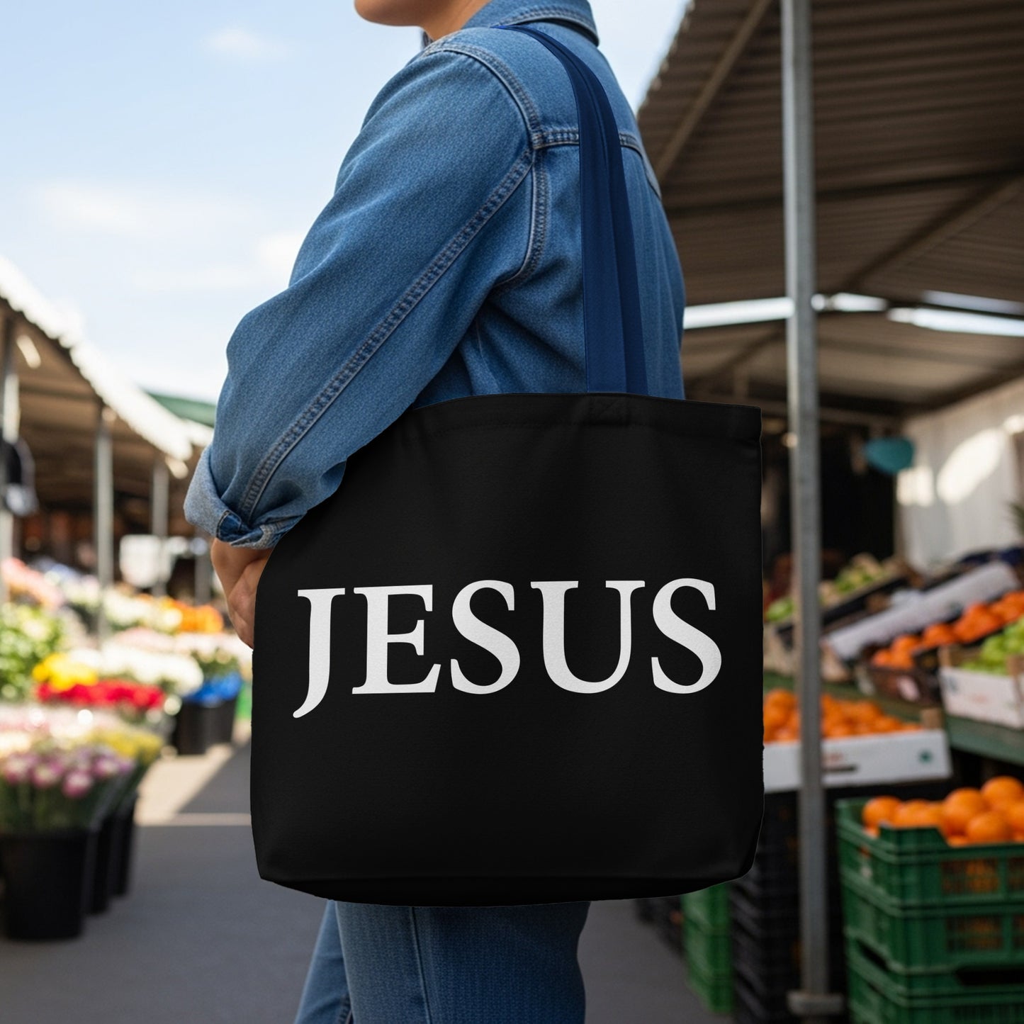 Black tote bag with white text that reads "JESUS" worn by a person in a denim jacket.