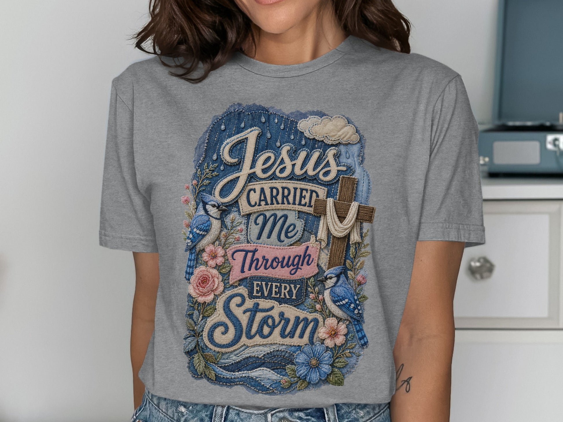 Grey t-shirt with "Jesus Carried Me Through Every Storm" text and floral cross design.