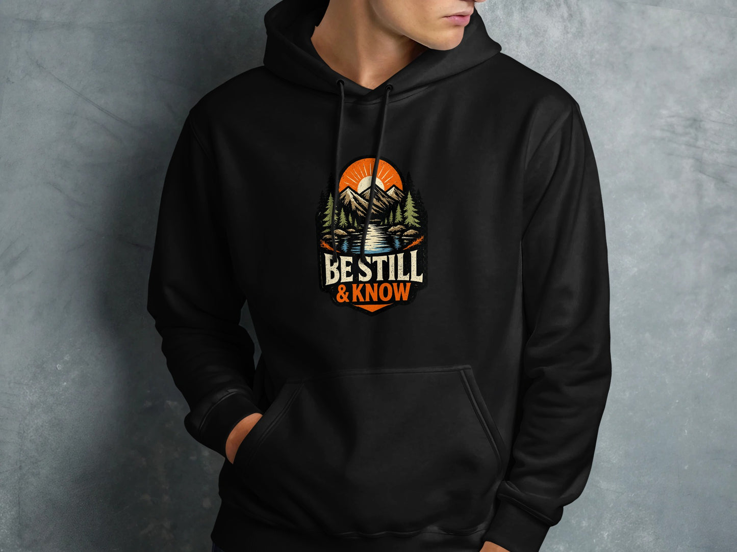 Black hoodie with a circular nature scene logo featuring mountains and text "BE STILL & KNOW".