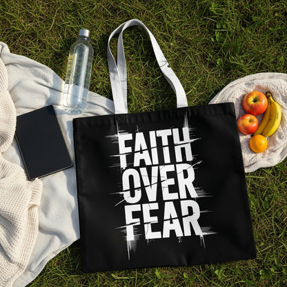 Black tote bag with white "FAITH OVER FEAR" text, laid on grass with fruit and water bottle.