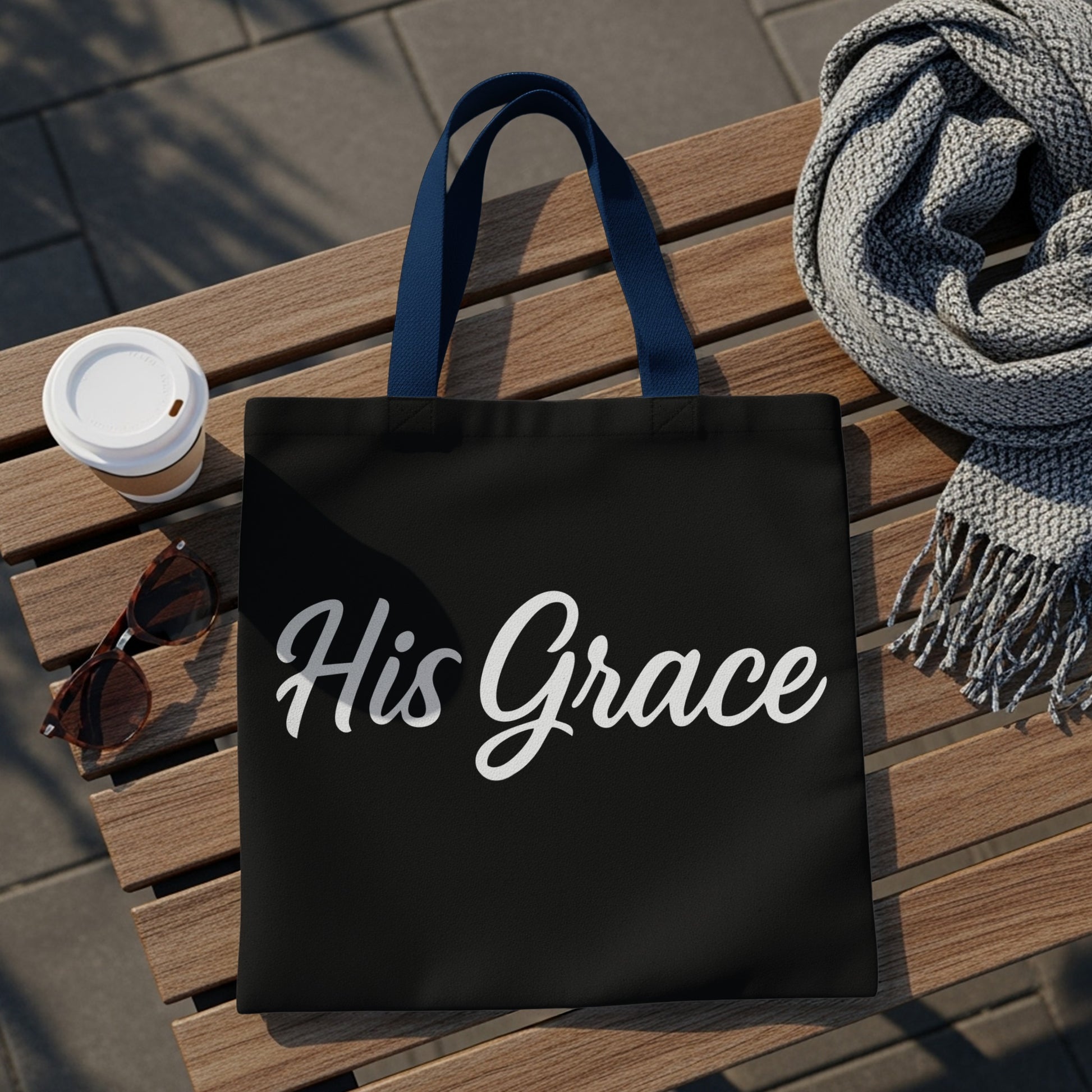 Black tote bag with "His Grace" in white script, on a wooden bench.