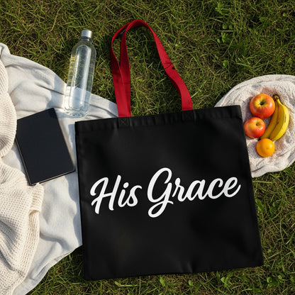 Black tote bag with white "His Grace" text and red handles on grass.