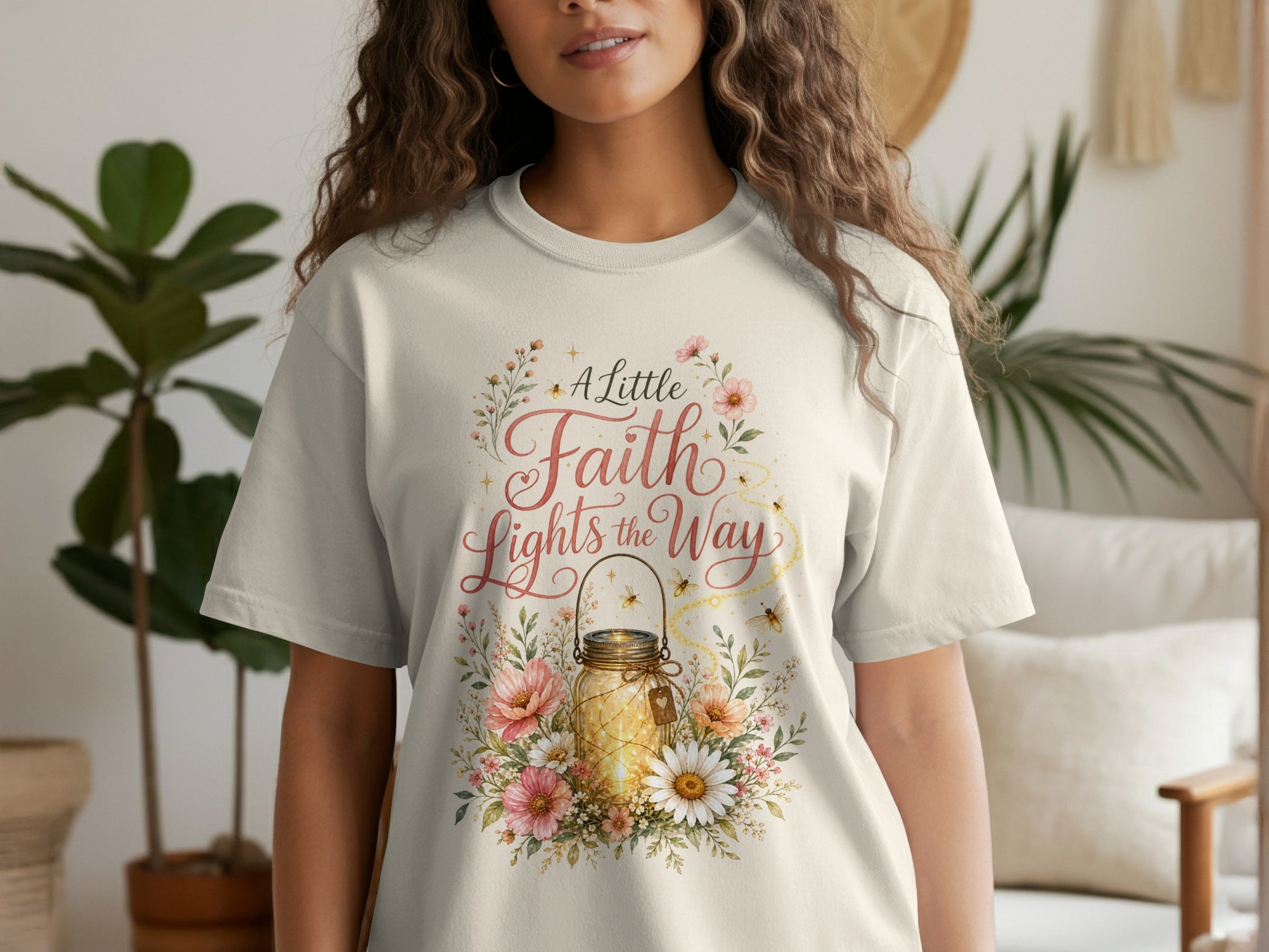 Woman wearing light cream t-shirt with "A Little Faith Lights the Way" text and a mason jar with lights.