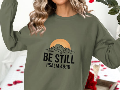 Olive green sweatshirt with mountains, sun, and text "BE STILL PSALM 46:10".