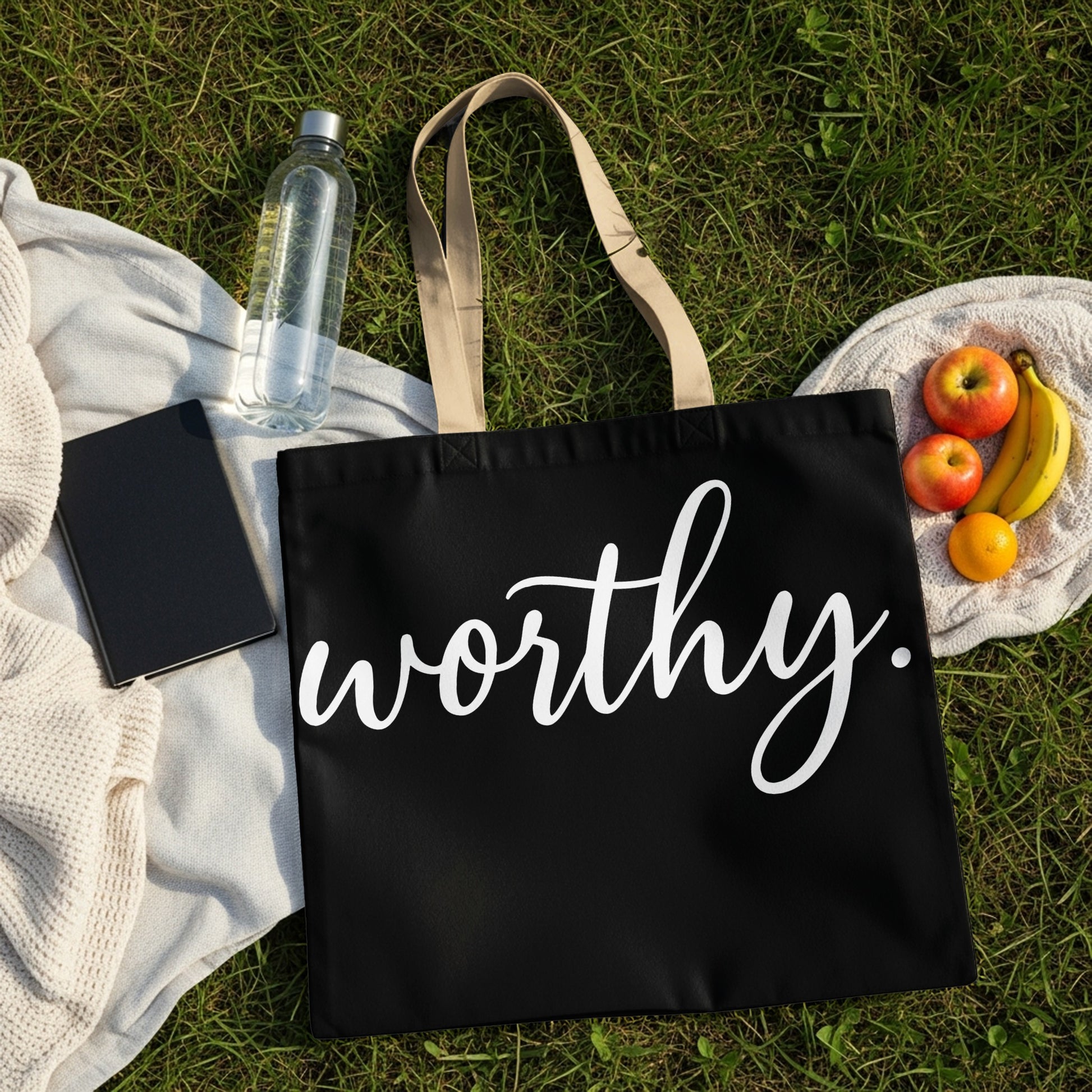Black tote bag with "worthy." in white script, beside fruit and a water bottle.