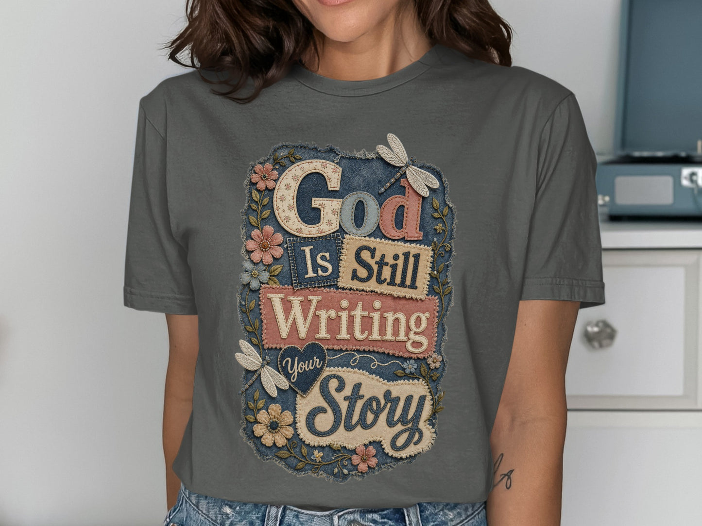 Grey t-shirt with "God Is Still Writing Your Story" text and floral embellishments.