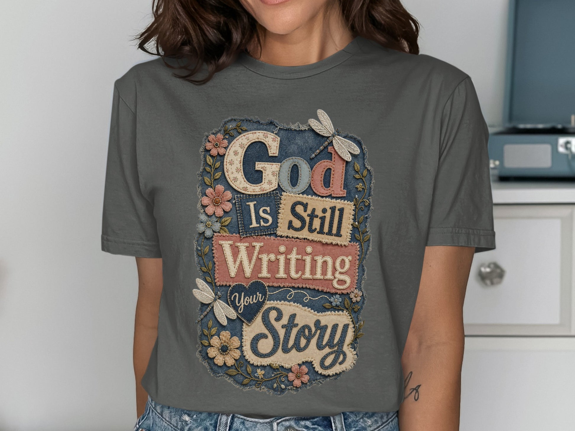 Grey t-shirt with "God Is Still Writing Your Story" text and floral embellishments.