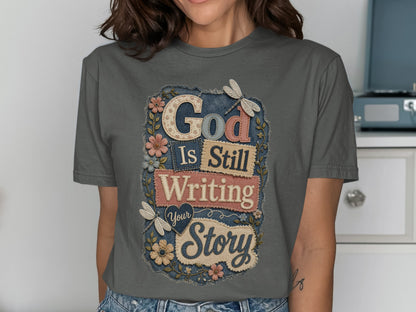 Grey t-shirt with "God Is Still Writing Your Story" text and floral embellishments.