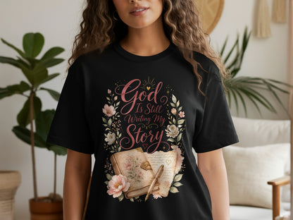 Black t-shirt with "God Is Still Writing My Story" graphic featuring floral wreath and open book.