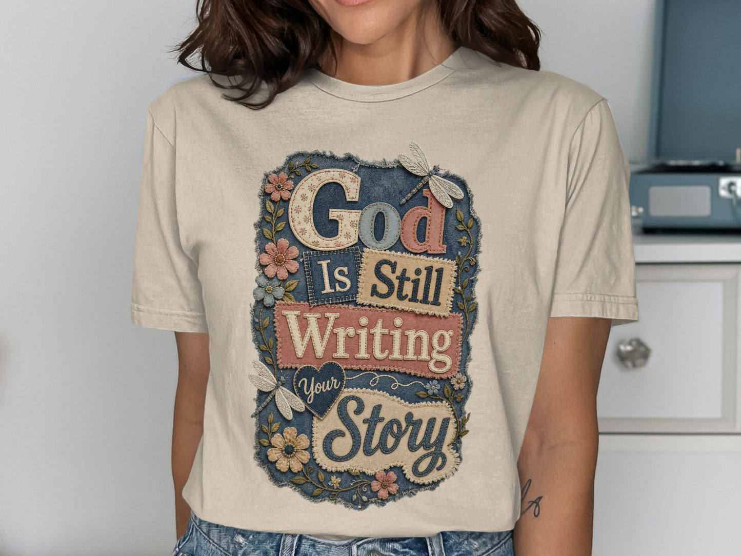 T-shirt with text "God Is Still Writing Your Story" and floral accents.