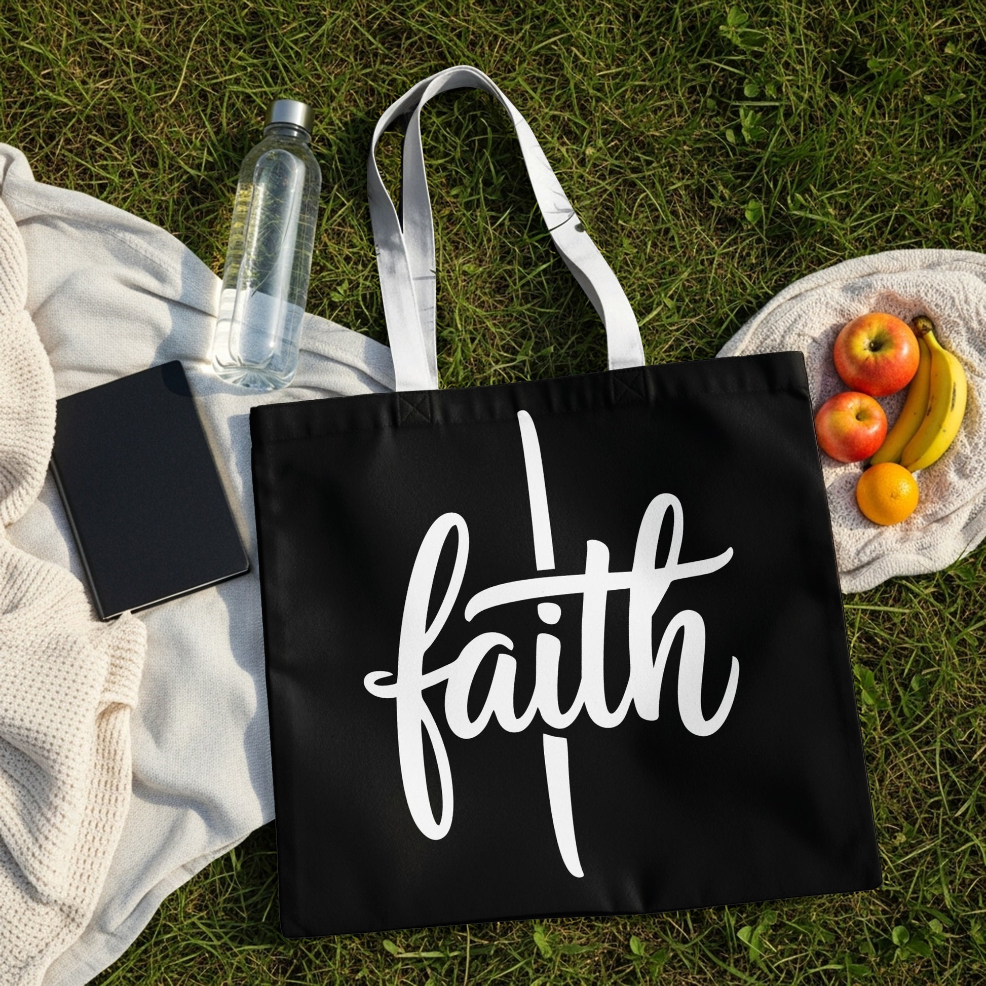 Black tote bag with white script lettering spelling "faith" on grass.