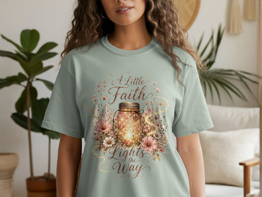 Light green t-shirt with a graphic of a mason jar filled with lights, surrounded by flowers and the text "A Little Faith Lights the Way."