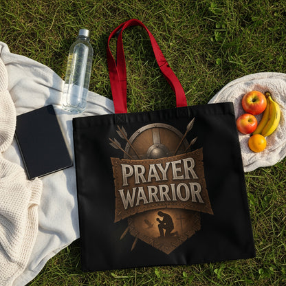 Black tote bag with "Prayer Warrior" text and shield design on grass.