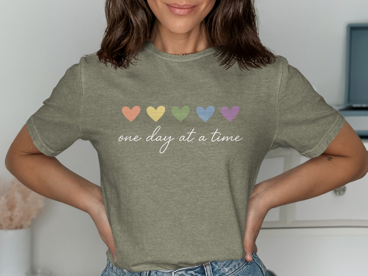Olive green t-shirt with five pastel hearts and "one day at a time" text.