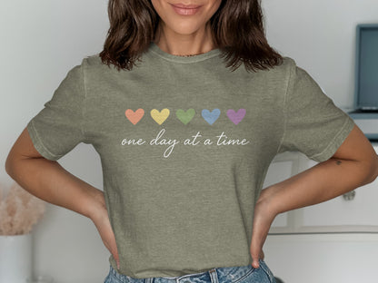 Olive green t-shirt with five pastel hearts and "one day at a time" text.
