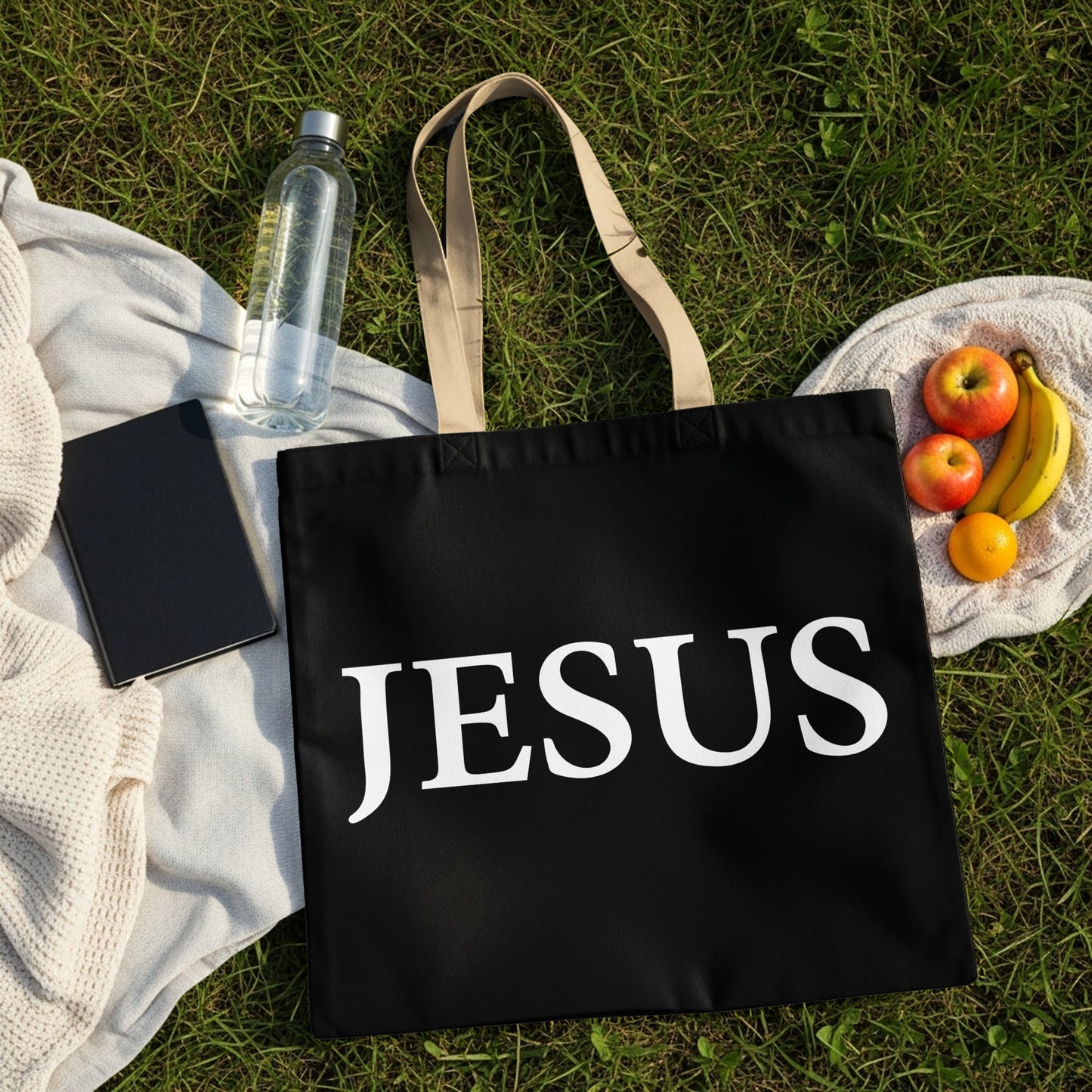 Black tote bag with "JESUS" printed in white, on a grassy background.