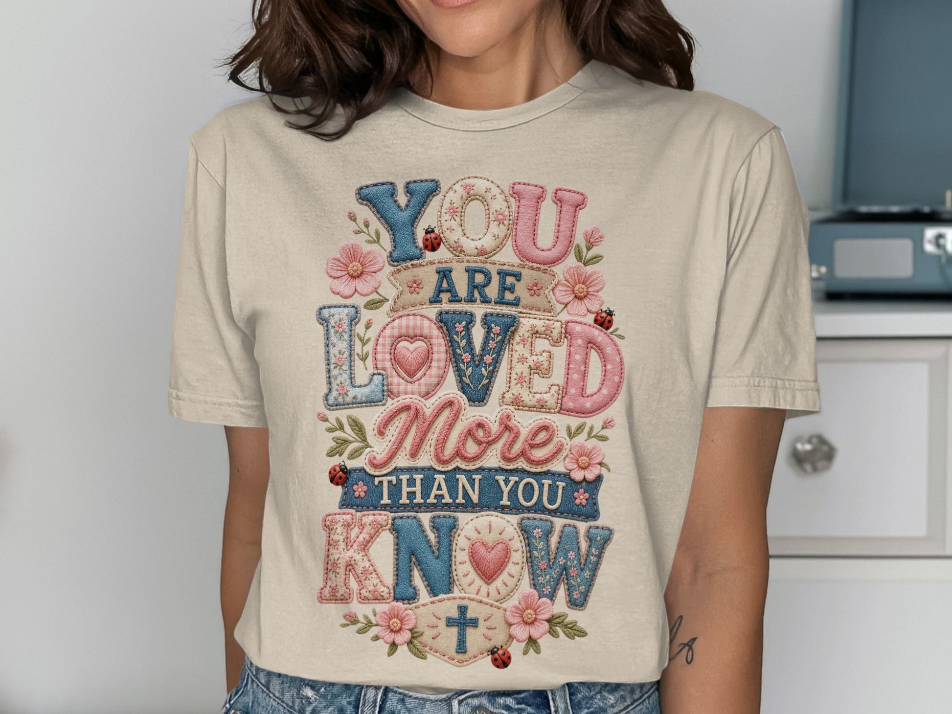 T-shirt with "YOU ARE LOVED MORE THAN YOU KNOW" text, decorated with flowers and ladybugs.