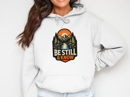 White hoodie with "Be Still & Know" text and mountain landscape design.