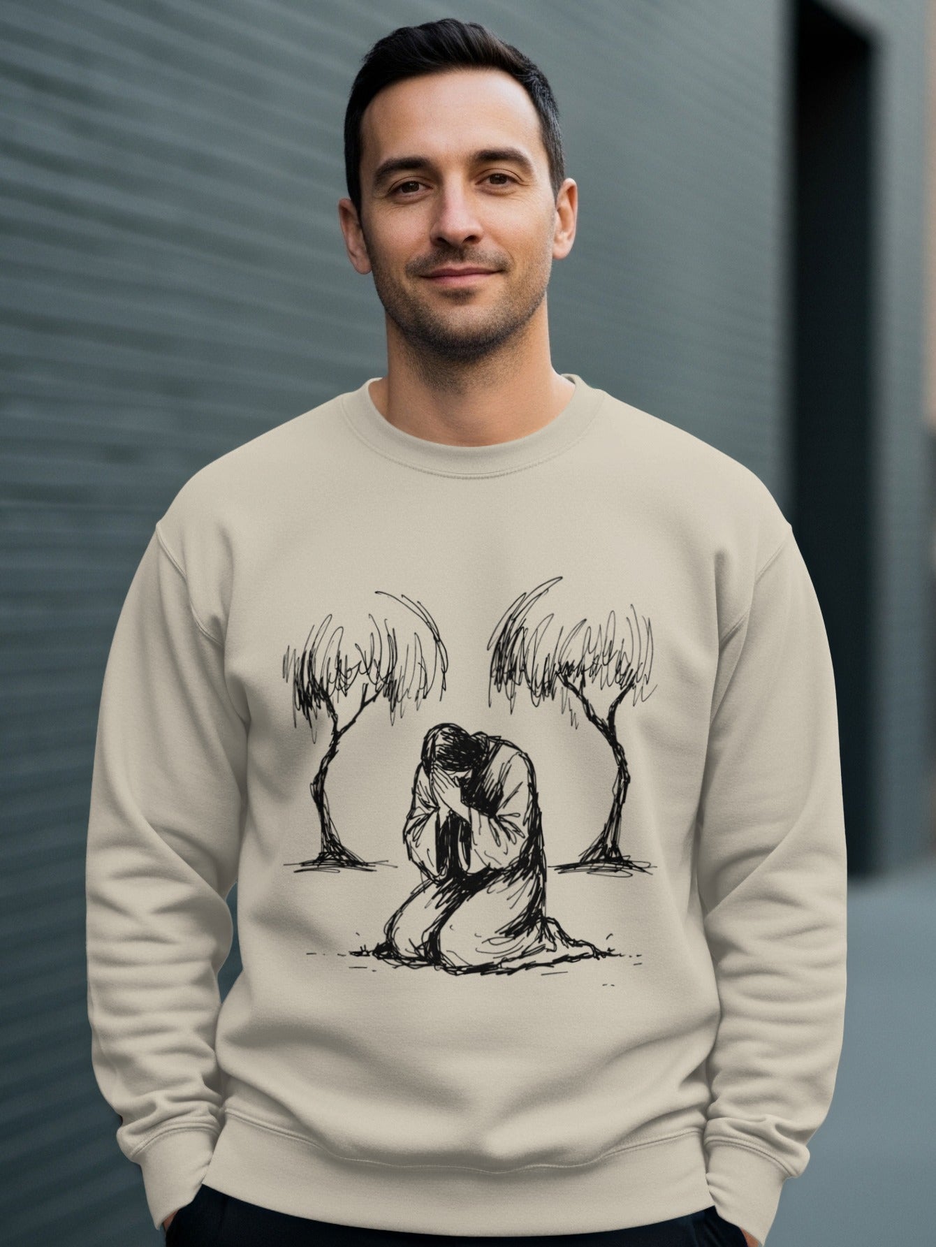 Man wearing a beige sweatshirt with a sketch of Jesus kneeling between two trees.