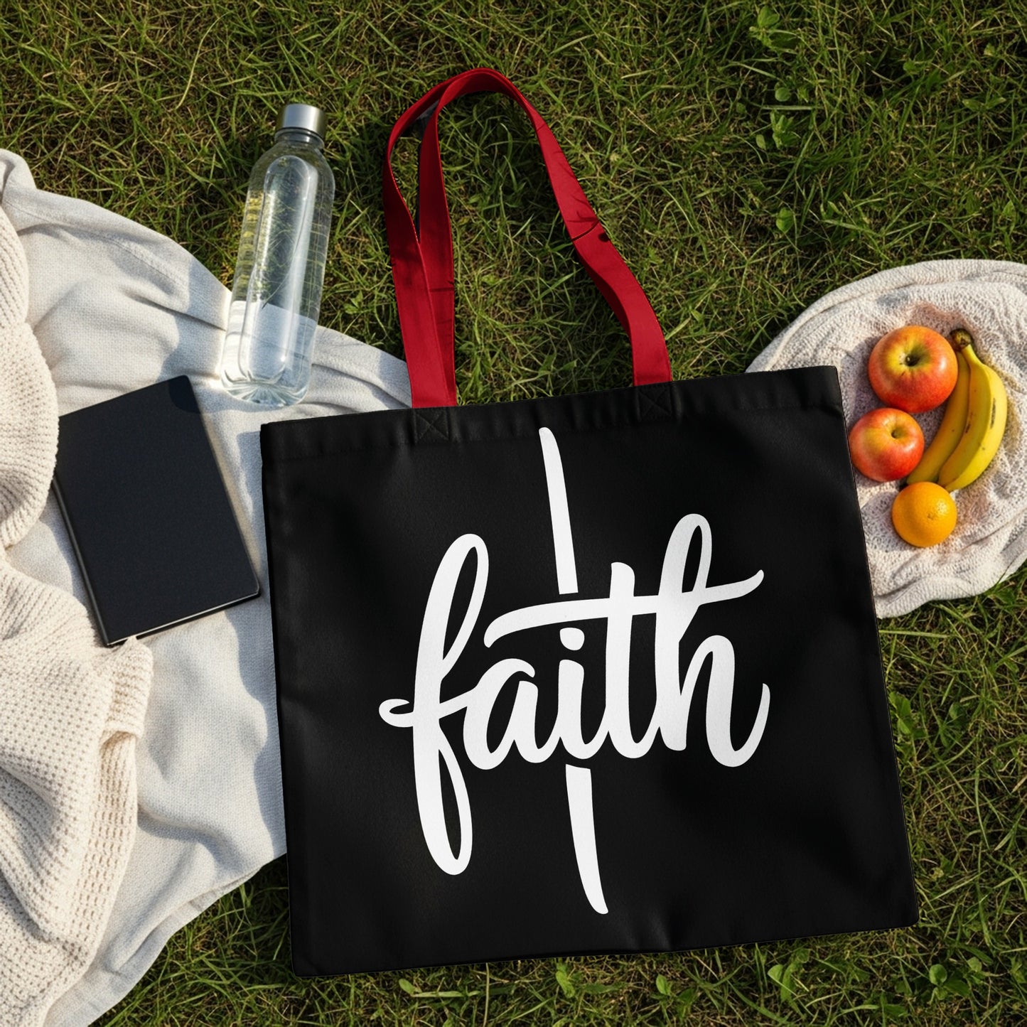 Black tote bag with white "faith" text and red handles, on grass with fruit and water.