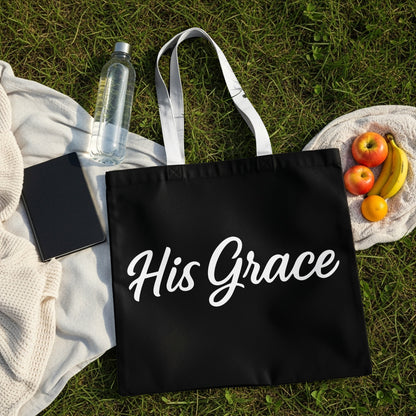 Black tote bag with white script text "His Grace" on grass with fruit and water bottle.