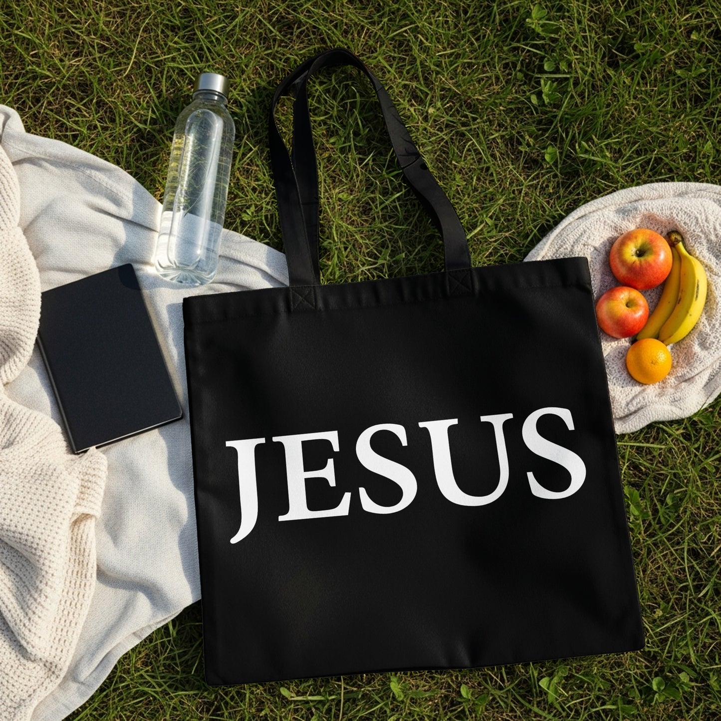 Black tote bag with "JESUS" printed in white, on grass with fruit and water.