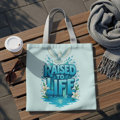 Light blue tote bag with "Raised to Life" text, a dove, and lilies.