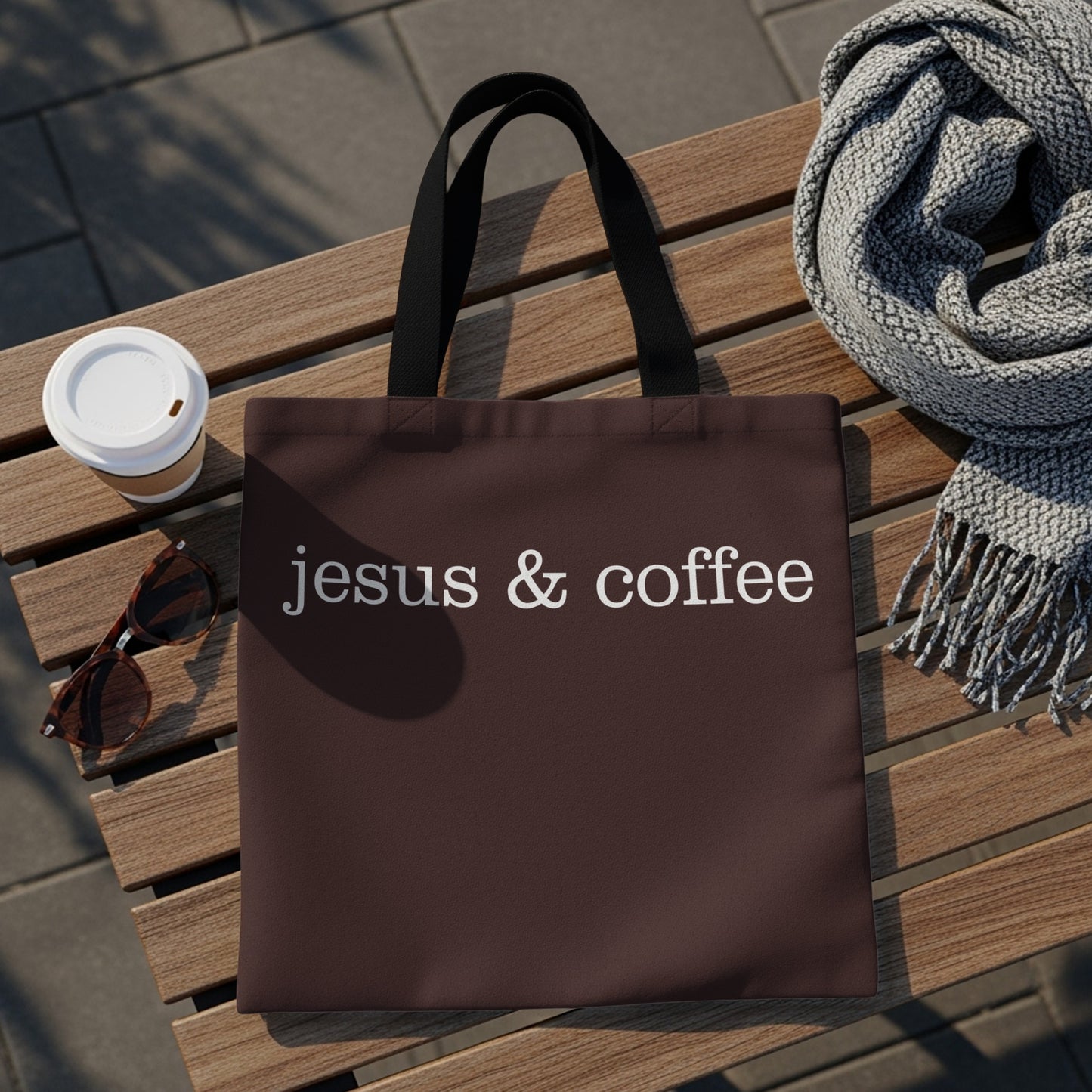 Brown tote bag with "jesus & coffee" text, coffee cup, and sunglasses on a bench.