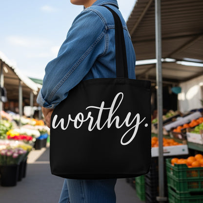 Black tote bag with "worthy." in white cursive text, held by person in denim jacket.